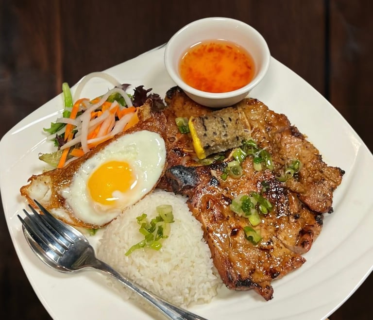Grilled meat with fried egg, rice, stir-fried vegetables, and red sauce on a white plate with fork