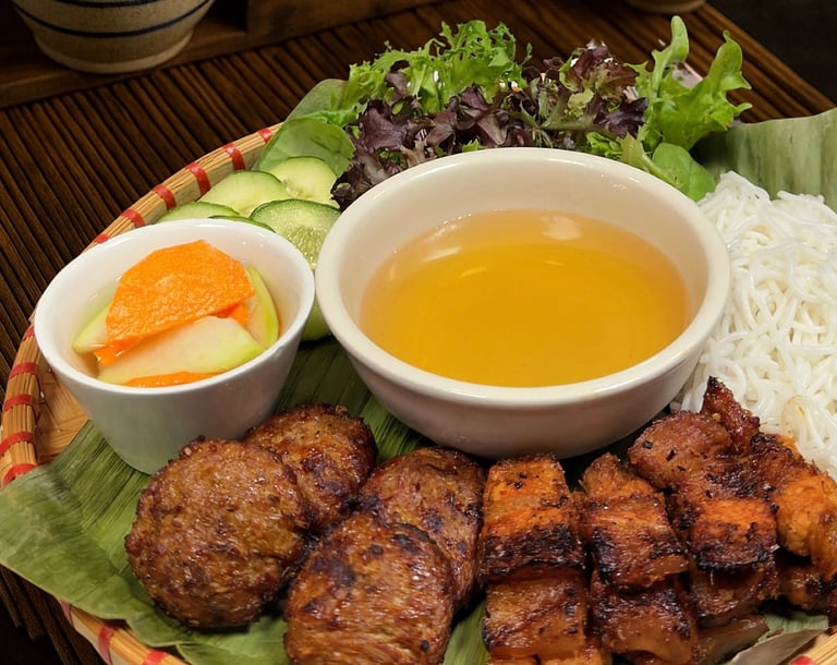 Asian food platter with fried meatballs, tofu cubes, fresh vegetables, rice noodles, and dipping sauce on banana leaf