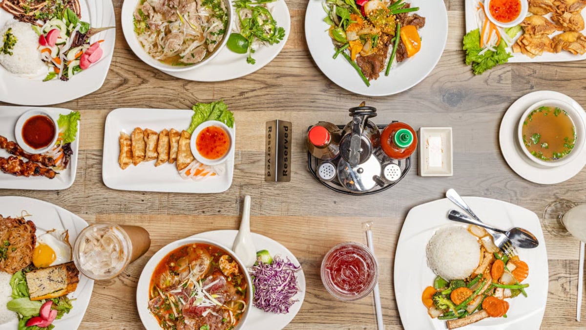 Overhead view of a wooden table with multiple plated Asian dishes, rice bowls, dipping sauces, and utensils arranged for a group meal